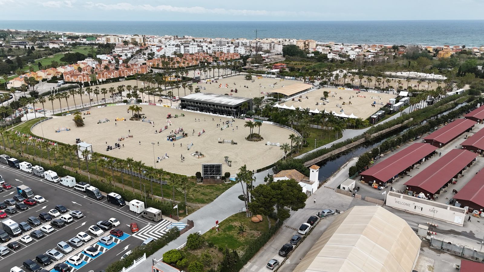 Vista aérea del recinto MET Oliva: varias pistas de arena Risohorse® con obstáculos de salto, el pueblo y el Mediterráneo al fondo